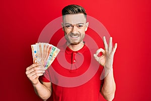 Young redhead man holding philippine peso banknotes doing ok sign with fingers, smiling friendly gesturing excellent symbol