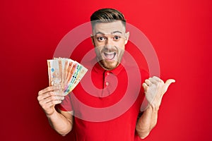 Young redhead man holding philippine peso banknotes pointing thumb up to the side smiling happy with open mouth