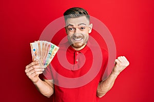 Young redhead man holding philippine peso banknotes screaming proud, celebrating victory and success very excited with raised arm