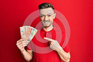 Young redhead man holding philippine peso banknotes smiling happy pointing with hand and finger