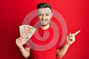 Young redhead man holding philippine peso banknotes smiling happy pointing with hand and finger to the side