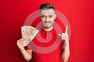 Young redhead man holding philippine peso banknotes smiling with an idea or question pointing finger with happy face, number one