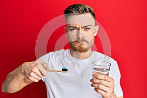 Young redhead man holding toothbrush with toothpaste and glass of water depressed and worry for distress, crying angry and afraid