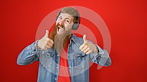 Young redhead man listening to music doing thumbs up gesture over isolated red background