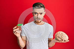 Young redhead man playing colorful puzzle cube intelligence game holding brain in shock face, looking skeptical and sarcastic,