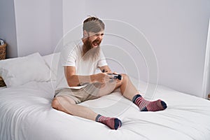 Young redhead man playing video game sitting on bed at bedroom