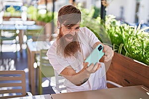 Young redhead man playing video game sitting on table at coffee shop terrace