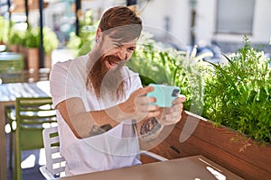 Young redhead man playing video game sitting on table at coffee shop terrace