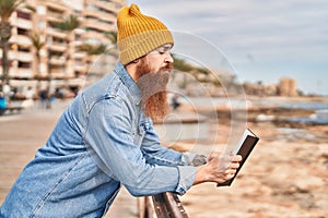 Young redhead man reading book with serious expression at seaside