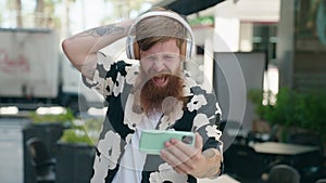 Young redhead man smiling confident watching video on smartphone at coffee shop terrace