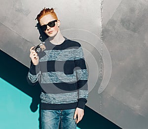 Young redhead man in a sweater and jeans standing next to green grea wall and taking photos vintage camera warm summer sunny day