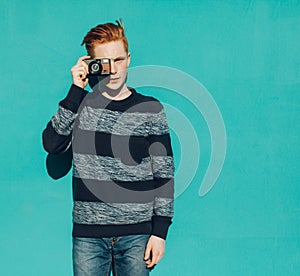 Young redhead man in a sweater and jeans standing next to turquoise wall and taking photos vintage camera warm summer sunny day