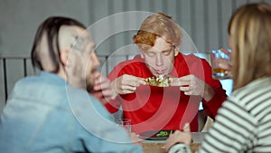 Young redhead man tasting delicious pizza in cafe and showing thumb up talking to friends sitting at table. Portrait of