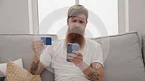 Young redhead man using smartphone and credit card sitting on sofa at home