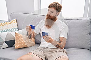 Young redhead man using smartphone and credit card sitting on sofa at home