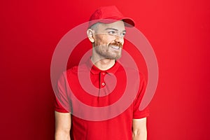 Young redhead man wearing delivery uniform and cap looking away to side with smile on face, natural expression