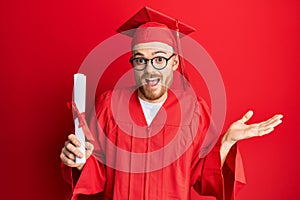 Young redhead man wearing graduation cap and ceremony robe holding degree celebrating achievement with happy smile and winner