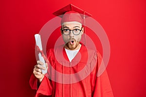 Young redhead man wearing graduation cap and ceremony robe holding degree scared and amazed with open mouth for surprise,