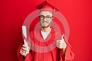 Young redhead man wearing graduation cap and ceremony robe holding degree smiling happy and positive, thumb up doing excellent and