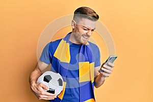 Young redhead man wearing hooligan scarf and using smartphone smiling and laughing hard out loud because funny crazy joke