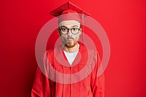 Young redhead man wearing red graduation cap and ceremony robe making fish face with lips, crazy and comical gesture