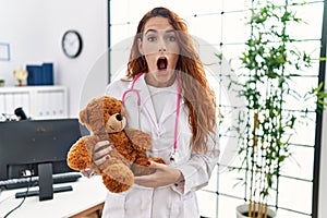 Young redhead pedriatician woman holding teddy bear scared and amazed with open mouth for surprise, disbelief face