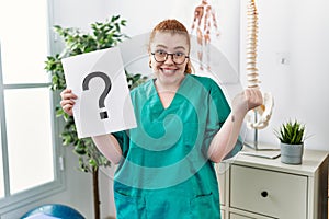 Young redhead physiotherapist woman holding question mark at the clinic screaming proud, celebrating victory and success very