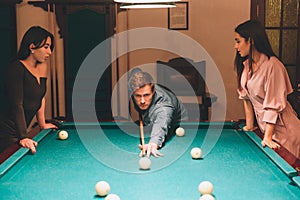 Young redhead player stand between two models. He aims into billiard ball. Young women stand and look at gamel. They lean to tabl