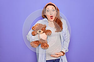 Young redhead pregnant woman expecting baby holding teddy bear over purple background scared in shock with a surprise face, afraid