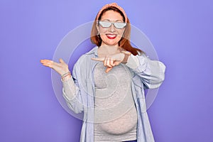 Young redhead pregnant woman expecting baby wearing funny thug life sunglasses amazed and smiling to the camera while presenting