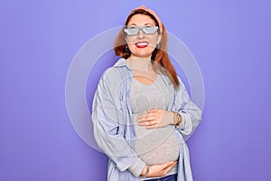 Young redhead pregnant woman expecting baby wearing funny thug life sunglasses looking away to side with smile on face, natural