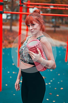 Young redhead sportswoman holding a foam roller and smiling in the park at vibrant autumn