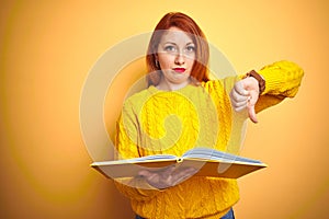 Young redhead student woman reading book standing over yellow isolated background with angry face, negative sign showing dislike