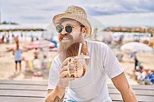 Young redhead tourist man drinking cocktail sitting on the bench at the beach