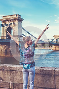 Young redhead woman arms raised and looking away at Chain Bridge, Budapest