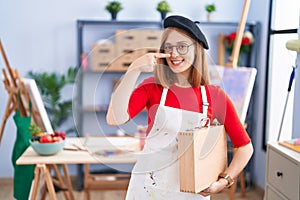 Young redhead woman at art studio holding art case pointing with hand finger to face and nose, smiling cheerful