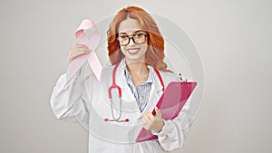 Young redhead woman doctor holding breast cancer awareness pink ribbon and clipboard over isolated white background