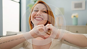 Young redhead woman doing heart symbol with hands sitting on sofa at home
