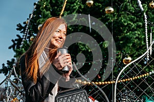Young redhead woman drinking coffee wandering near Christmas tree in the streets on winter city. Happy young smiling