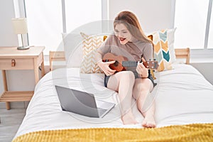 Young redhead woman having online ukulele lesson sitting on bed at bedroom