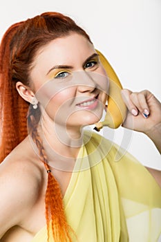 Young redhead woman holding banana near her face like smartphone on a white background.