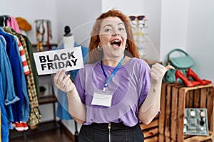 Young redhead woman holding black friday banner at retail shop pointing thumb up to the side smiling happy with open mouth