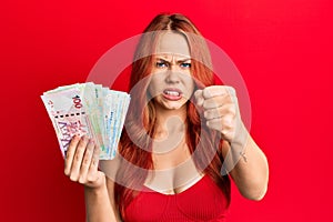 Young redhead woman holding hong kong dollars banknotes annoyed and frustrated shouting with anger, yelling crazy with anger and
