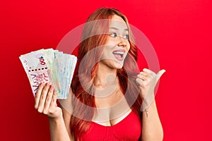 Young redhead woman holding hong kong dollars banknotes pointing thumb up to the side smiling happy with open mouth