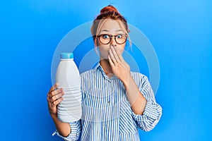Young redhead woman holding liter bottle of milk covering mouth with hand, shocked and afraid for mistake