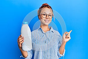 Young redhead woman holding liter bottle of milk smiling happy pointing with hand and finger to the side