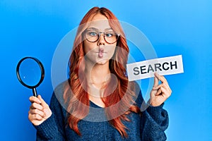 Young redhead woman holding magnifying glass and search word making fish face with mouth and squinting eyes, crazy and comical