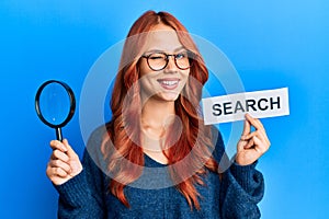 Young redhead woman holding magnifying glass and search word winking looking at the camera with sexy expression, cheerful and