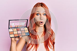 Young redhead woman holding makeup nudes scared and amazed with open mouth for surprise, disbelief face