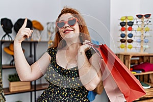 Young redhead woman holding shopping bags at retail shop smiling with an idea or question pointing finger with happy face, number